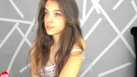 Media: Video of a young woman with long brown hair, light skin, and brown eyes, wearing a white tank top, sitting against a geometric-patterned gray wall.