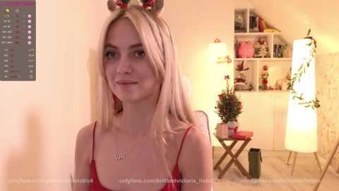 Media: Video of a smiling, blonde woman in a red dress with mouse ears, standing in a warmly lit, cozy room with shelves, a lamp, and a potted plant.