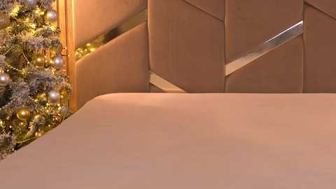 Media: Video of a cozy bedroom with a beige tufted headboard, a warm golden Christmas tree adorned with white and gold ornaments on the left, and a neatly made bed covered in soft, beige bedding.