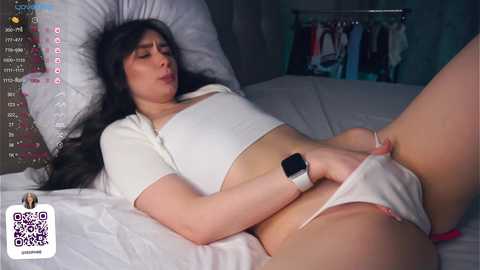 Media: A video of a woman with long black hair, wearing a white crop top and panties, lying on a bed, masturbating. The background shows a closet with clothes.