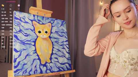 Media: A young Asian woman with fair skin and brown hair in a ponytail, wearing a light pink cardigan over a cream lace top, stands next to a painted canvas of a sad yellow cat with blue waves.