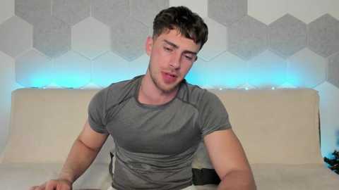 Media: Video of a young, fit man with short, dark hair, wearing a tight, gray athletic shirt, sitting on a bed with a beige headboard. The background features a hexagonal, gray and white patterned wall.