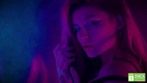 Media: Video of a woman with long, blonde hair, wearing a black top, under a dim, neon-lit setting. She's holding her hand to her face, with a sensual expression.