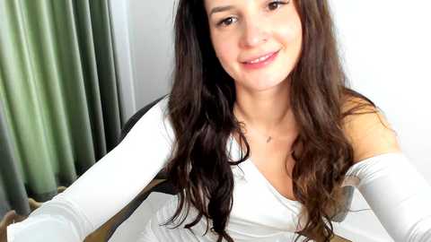 Media: Video of a smiling young woman with long, wavy dark brown hair, fair skin, and a medium build. She wears a white off-shoulder top and sits on a black chair in a brightly lit room with green curtains.