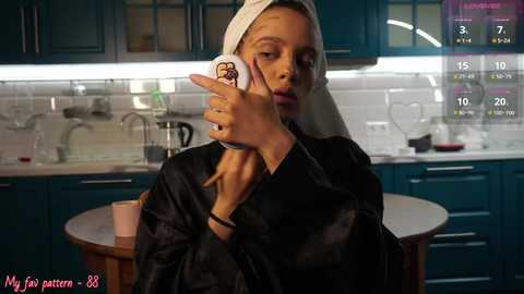 Media: A video shows a woman with a towel on her head, holding a phone in a modern kitchen. She wears a black robe. Background includes blue cabinets, a sink, and a calendar.