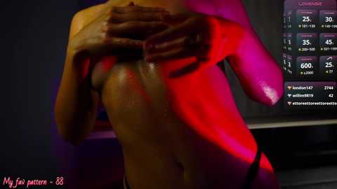 Media: Video of a nude woman with medium skin tone, standing in a dimly-lit room, hands covering her chest. Background shows a digital screen displaying financial charts with red and pink hues.