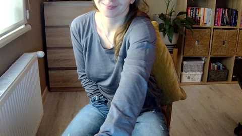 Media: A video of a smiling woman with light skin, long brown hair, wearing a blue long-sleeve shirt and jeans, sitting on a wooden chair in a cozy, organized living room with beige walls, a radiator, bookshelf, and wicker baskets.