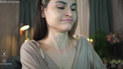 Media: Video of a fair-skinned woman with long, straight dark hair, wearing a light pink blouse, eyes closed, smiling softly, in a dimly lit room with blurred green curtains and a table with flowers in the background.