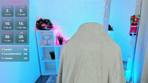 Media: Video of a white-tiled room with a beige towel-draped massage table, illuminated by soft blue lights. The room has a modern, minimalist design with a shelf holding a small potted plant and a mirror.