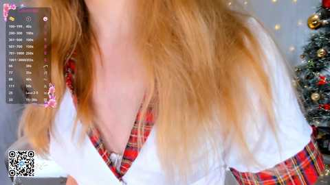 Media: Video of a woman with long, wavy blonde hair, wearing a white top with red plaid accents, against a blurred Christmas tree background.