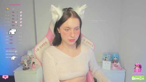 Media: Video of a pale-skinned woman with shoulder-length dark hair wearing a white cat headband and cropped sweater, seated in a pink gaming chair, surrounded by anime figurines in a pastel-colored room.