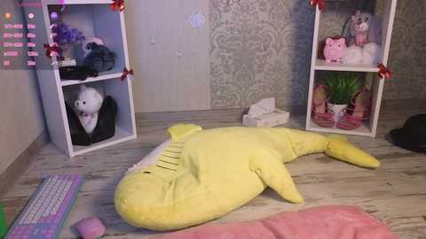 Video of a cozy bedroom with white walls, a plush yellow whale toy, two white shelves adorned with stuffed animals and a plush owl, and a pink blanket on the floor.