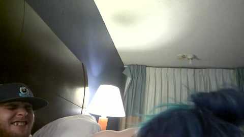 Media: Video of a man in a black hat and a woman with blue hair in a bed, with a lamp emitting a soft light and a white curtain in the background.