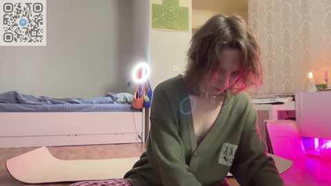 Media: A video of a young woman with messy hair in a green robe, sitting on a pink mat in a dimly-lit room with a bed, wall art, and a candle.