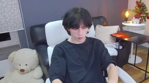 Media: Video of a young man with short black hair, wearing a black T-shirt, sitting in a white leather armchair in a cozy, dimly lit room with a beige teddy bear, a small round table with a lamp, and a floral arrangement on the right.