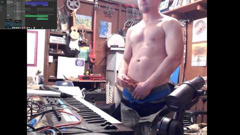 Media: A video of a shirtless, light-skinned man with a muscular build, wearing blue shorts, standing in a cluttered, wooden-walled room filled with musical equipment and instruments.