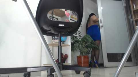 Media: Video of a woman in a blue dress and red heels standing near a black office chair, with a mirror reflecting her. Background features a potted plant, shelves with books, and a white door.