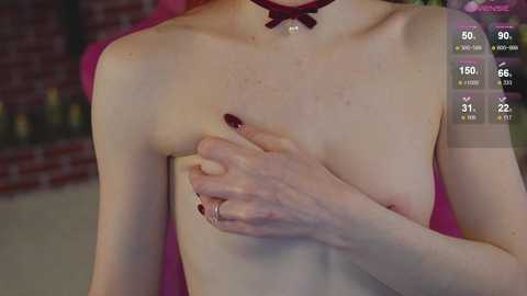 Media: A video of a fair-skinned, slender woman with red hair, topless, covering one small breast with her hand, wearing a red choker and ring, in a brick-walled room with pink furniture.