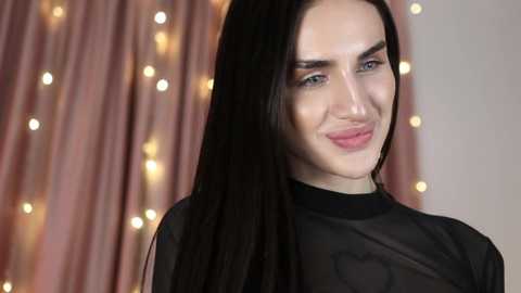 Media: Video of a young, fair-skinned woman with long, straight black hair, wearing a sheer black top. She has a confident smile and blue eyes. Background features soft, pink curtains with warm, bokeh lights.