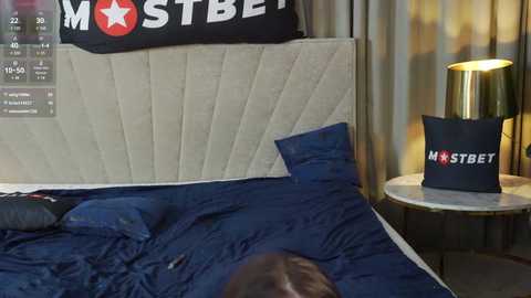 Media: Video of a modern bedroom with a beige upholstered headboard, navy blue bedding, a black \"Mostbet\" pillow, and a small wooden nightstand with a gold lamp.