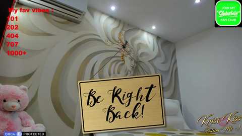 Media: A video of a modern bedroom with a beige and white abstract wall mural. A plush pink teddy bear sits on a bed. A wooden sign with the phrase \"Be Right Back!\" hangs in the center.