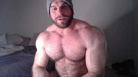 Media: Video of a muscular, bearded, pale-skinned man with a gray beanie, shirtless, sitting on a bed with rumpled gray sheets.