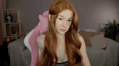 Media: Video of a young woman with long, straight red hair, pale skin, and a slim physique, wearing a black strapless top, sitting in a cozy, pink-themed room with plush toys, shelves, and a potted plant.