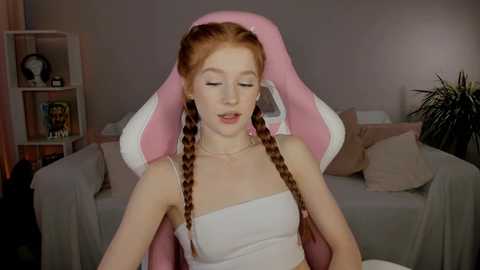 Media: Video of a young, fair-skinned woman with long red braids, wearing a white strapless top, seated in a pink gaming chair, surrounded by a cozy living room with soft pastel colors and a green plant.