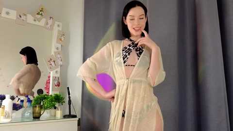 Media: Video of a slender, light-skinned Asian woman with shoulder-length black hair, wearing a sheer, beige robe and a black-and-white patterned bra. She stands confidently, adjusting her robe. Background features a white vanity with makeup and a mirror, and a gray curtain.