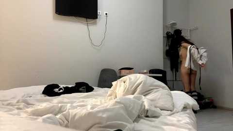 Media: Video of a messy bedroom with a white bed, black clothes, a TV, and a woman in a white dress and black shoes.