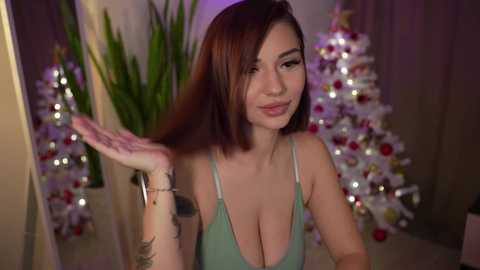 Media: Video of a smiling East Asian woman with shoulder-length brown hair, large breasts, and a green tank top, waving her hand, standing in front of two decorated Christmas trees in a dimly-lit room.