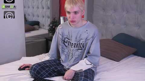 Media: Video of a young, slender, fair-skinned man with short, platinum blonde hair, wearing a grey graphic tee and plaid pajama pants, sitting on a bed in a modern bedroom.