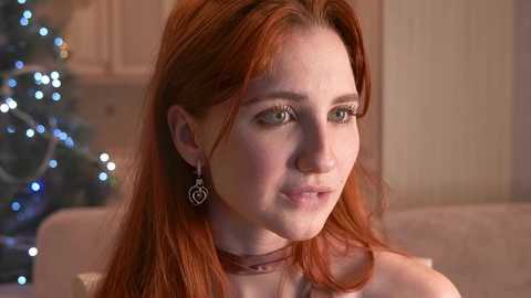 Media: Video of a fair-skinned woman with long red hair, wearing a choker and heart-shaped earrings, looking thoughtful in a softly lit room with a Christmas tree in the background.