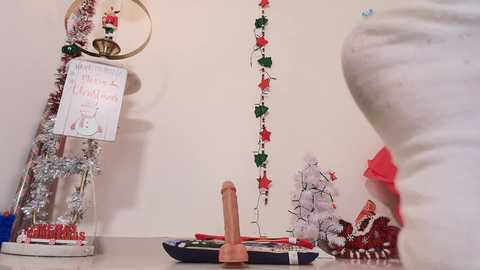 Media: A video of a festive room with a large, white mannequin wearing a red dress, decorated with Christmas garlands and a penis-shaped ornament on the table.