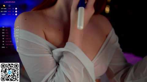 Media: A video of a woman with long, red hair, wearing a sheer, off-shoulder top, holding a white, cylindrical object to her lips. The background is dimly lit with blurred, indistinct shapes. QR code in the bottom left.