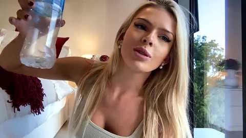 Media: Video of a blonde woman in a beige tank top, holding a water bottle, in a modern bedroom with white bedding and a window.