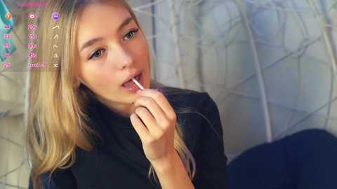 Media: Video of a young woman with long blonde hair, wearing a black top, holding a lollipop near her lips, against a blurred, white-tiled background with a pink chat box overlay.