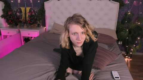 Media: Video of a Caucasian woman with shoulder-length, wavy blonde hair, wearing a black top, kneeling on a bed with a tufted headboard in a dimly lit room with purple and pink lighting.