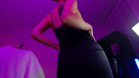 Media: A video of a woman in a tight black dress, illuminated by purple lighting, standing with her hands on her hips, in a dimly-lit room with a bed and a television.