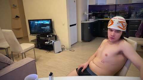 Media: Video of a young, fair-skinned, shirtless man in Star Wars-themed hat and shorts, sitting in a modern kitchen, watching TV.