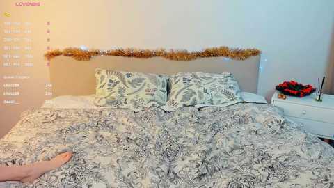 Media: Video of a bedroom with a white bed adorned with a patterned duvet, two pillows, and a garland of gold tinsel above the headboard. A person's bare feet are visible at the foot of the bed.