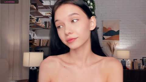 Video of a young Asian woman with long black hair, fair skin, and a slim build, wearing a floral headband, standing topless in a modern, minimalist room with white brick walls, bookshelves, and abstract artwork.