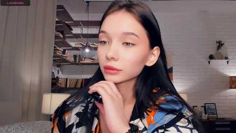 Video of an Asian woman with long black hair, light skin, and red lipstick, wearing a colorful patterned top, in a cozy, bookshelf-filled room with white brick walls.