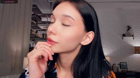 Video of a young woman with long black hair, closed eyes, and hand to her lips, in a modern, cozy room with white brick walls, shelves, and a lamp.