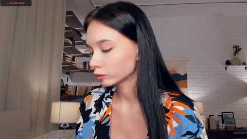 Video of a young Asian woman with long black hair, fair skin, and a slender build, wearing a colorful patterned blouse, standing in a modern, bright room with white brick walls and shelves filled with books and decor.