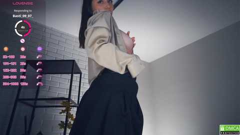 Video of a woman in a beige jacket and black skirt, standing in a modern room with white brick walls and a black metal shelf.