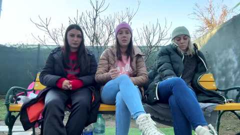 Media: Video of three women sitting on a park bench, dressed in winter clothes, with bare trees and a green fence in the background.