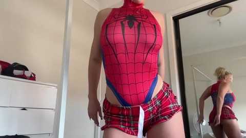 Media: Video of a woman in a red Spider-Man bodysuit, plaid mini skirt, and high-cut blue shorts. She stands in a bedroom with a white dresser, mirror, and beige walls.