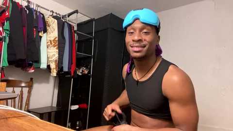 Media: Video of a muscular Black man with a blue headband, wearing a black sports bra, sitting in a small, cluttered room with clothes on racks and a wooden table.