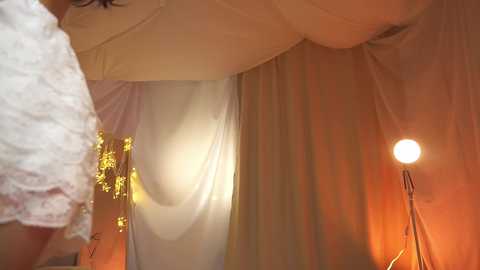 Media: Video of a person, likely a woman, wearing a sheer white dress with gold tassels, standing in a dimly lit room with an orange lamp and a white curtain.